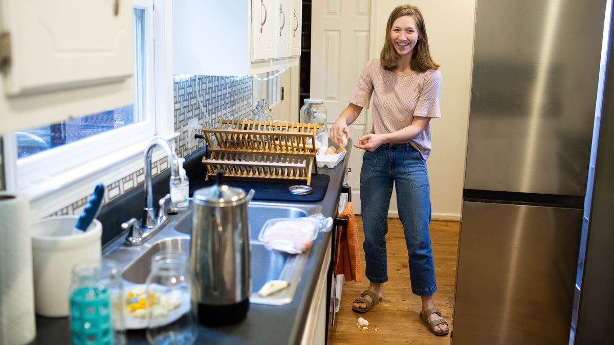 A woman smiling in a kitchen while holding a small white container next to a sink, with a wooden dish rack, coffee maker, and stainless steel refrigerator visible.